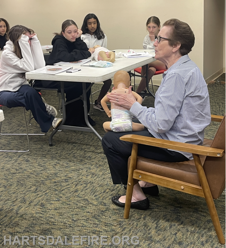 A woman teaches a group of children using a doll in a classroom setting, likely focusing on care or safety topics.
