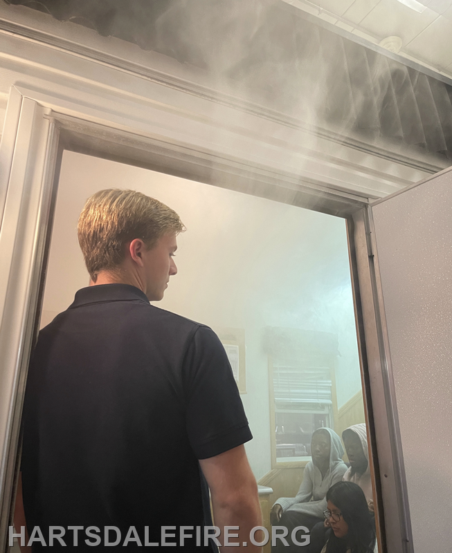A person stands at a doorway with smoke diffusing from the room, while others sit inside, likely in a training scenario.
