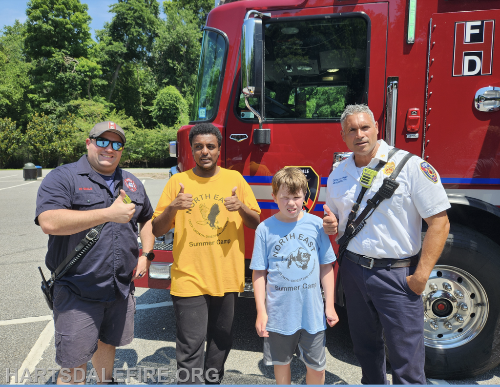 Three people pose in front of a fire truck, with two adults in uniforms and a child wearing a summer camp shirt.