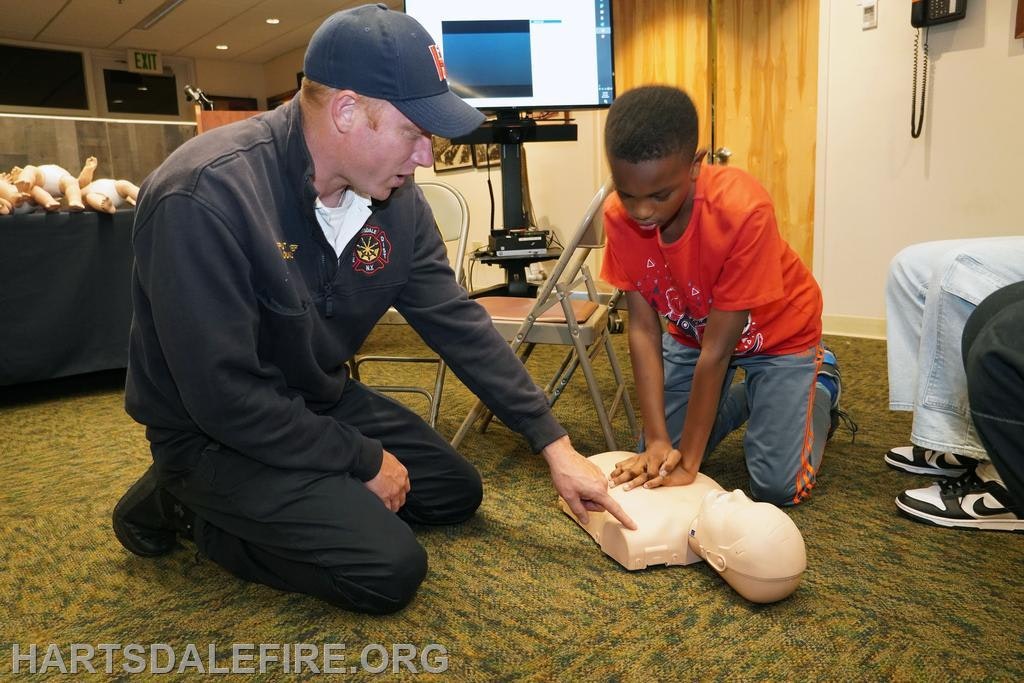 A child is learning CPR on a mannequin, guided by a firefighter in a classroom setting.
