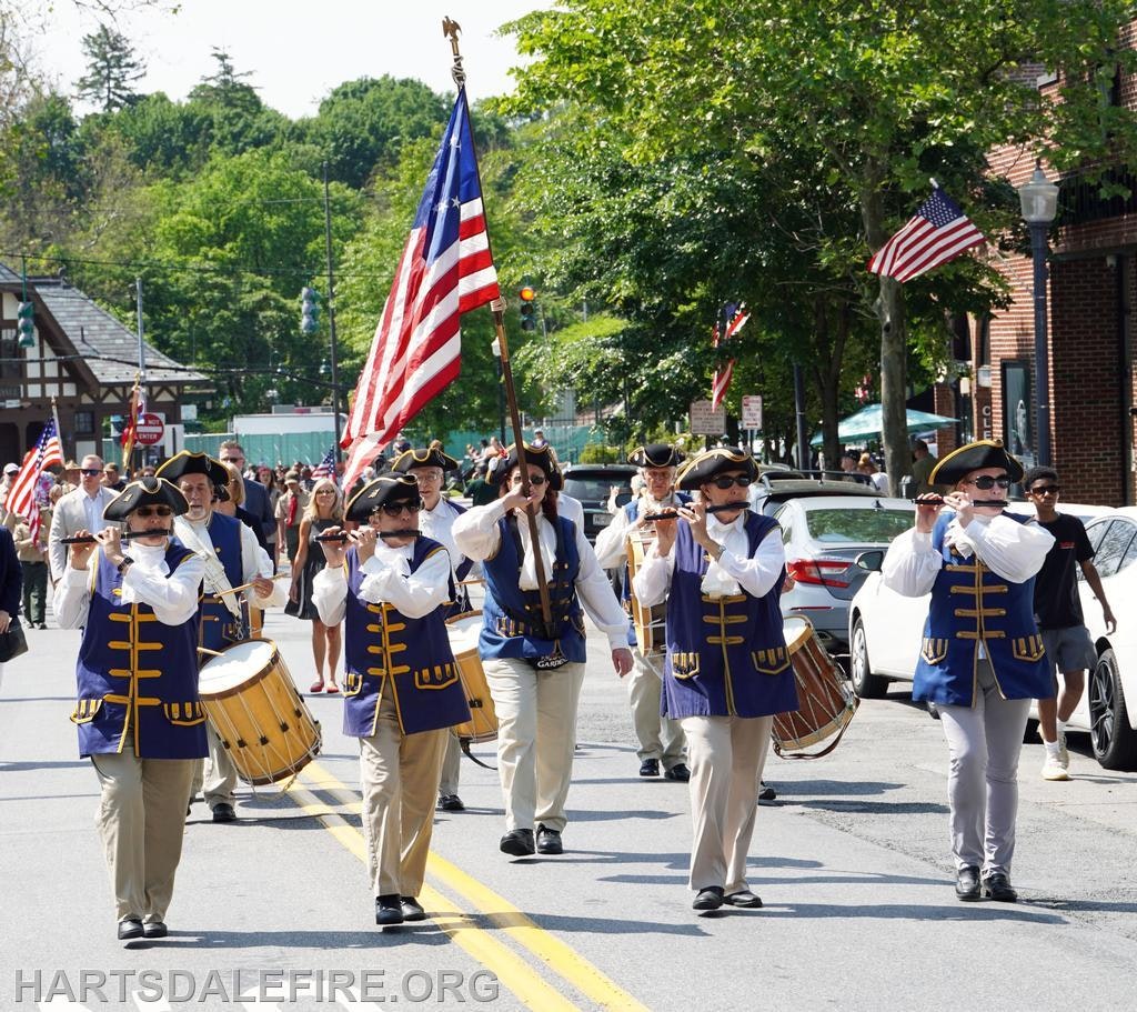A marching band with musicians in historical costumes plays and carries an American flag in a parade.