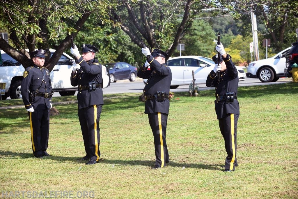 Four uniformed officers in a park, three holding rifles, performing a ceremonial salute.