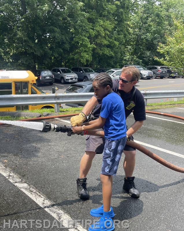A firefighter helps a child operate a fire hose, spraying water in a parking area surrounded by trees and vehicles.