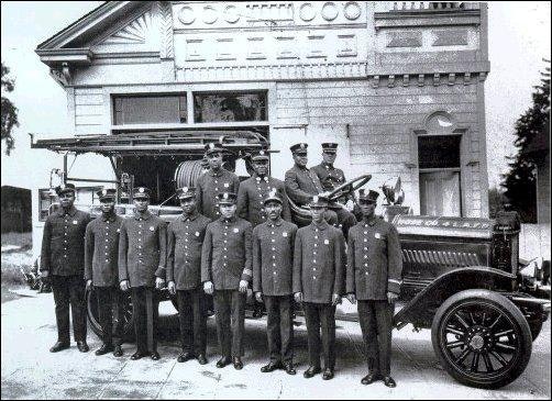 A group of uniformed firefighters stand in front of a vintage fire truck, with a station building in the background.