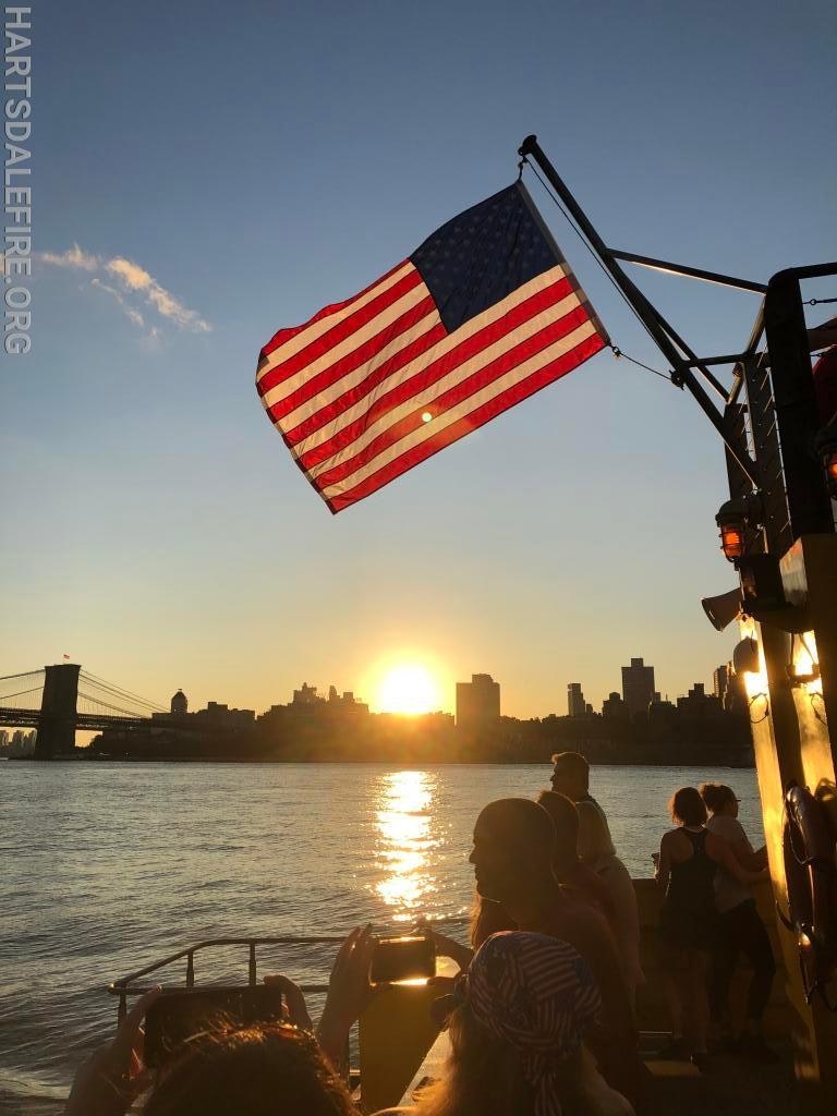 US flag on a boat at sunset, with a city skyline and a bridge in the background, surrounded by people.
