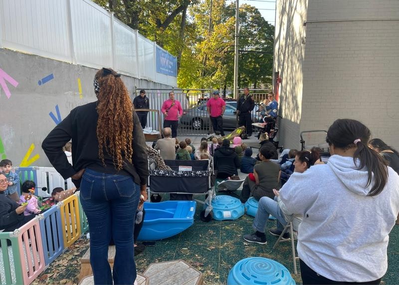 A crowd of adults and children sit outside, listening to speakers in pink shirts at a community event or gathering.