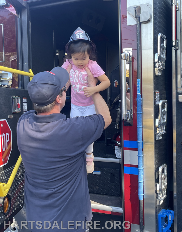 A firefighter helps a young girl wearing a firefighter hat step out of a fire truck.