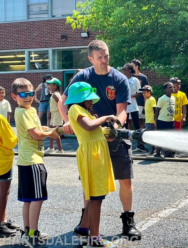 A firefighter helps kids spray water with a hose during an outdoor activity.
