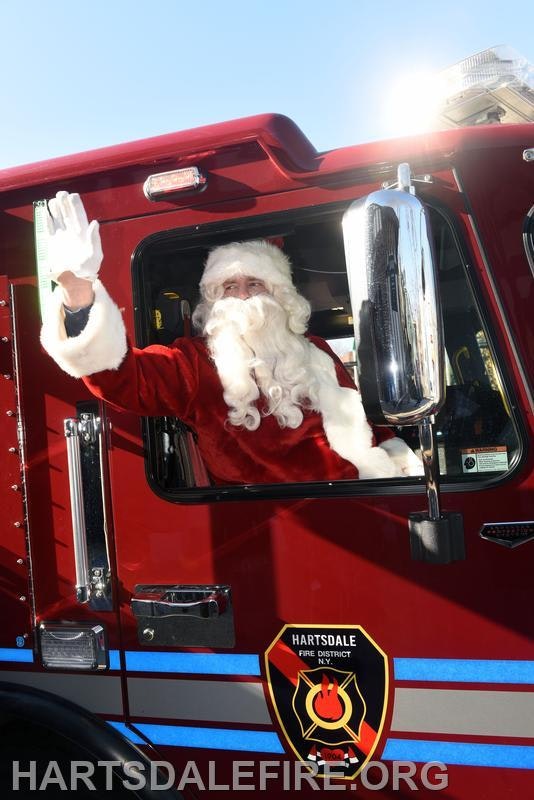 A man dressed as Santa waves from the window of a fire truck, representing a festive community event.
