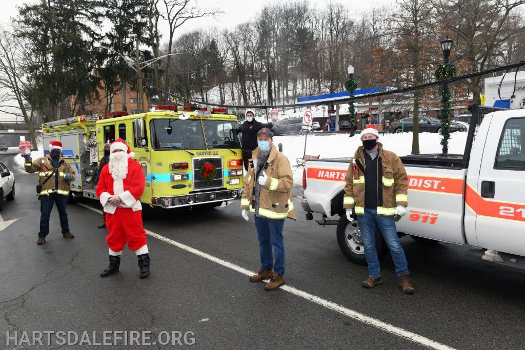 Santa and firefighters in festive gear in front of fire trucks on a snowy street.