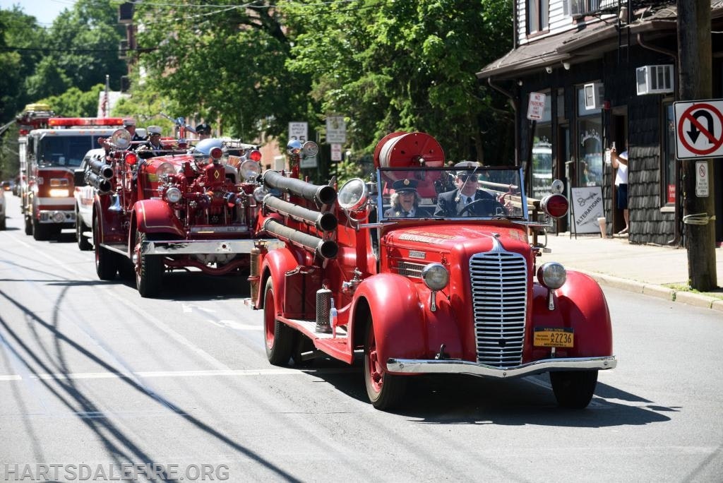 Vintage fire trucks driving in a parade with people on board, on a sunny day.