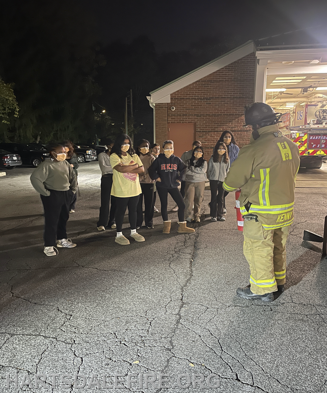 A firefighter is demonstrating safety techniques to a group of masked students at night outside a fire station.