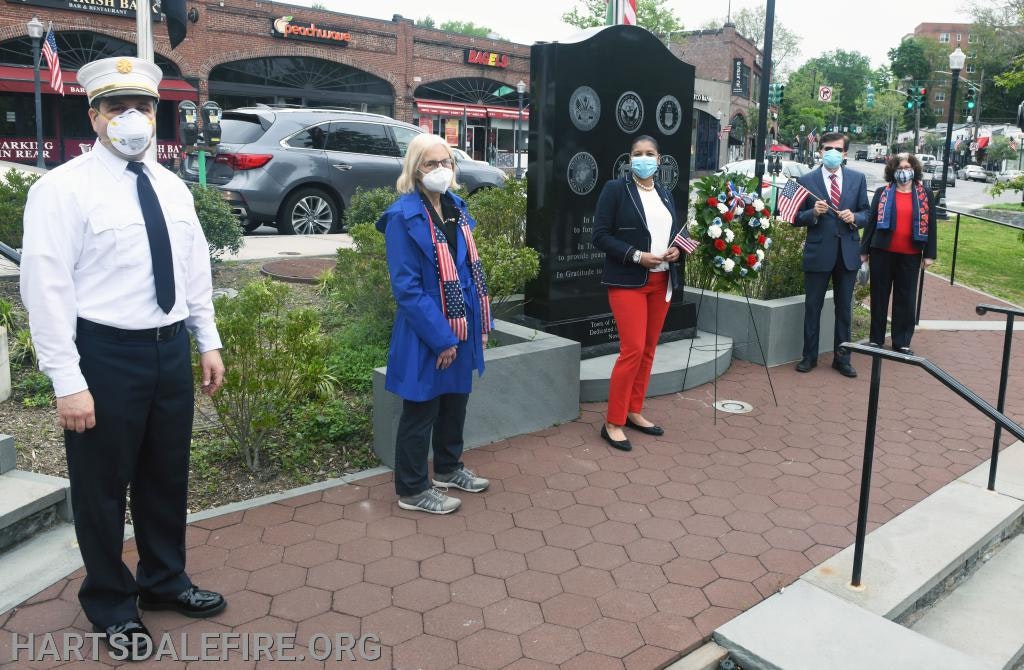 People in masks stand by a war memorial, holding flags, with a floral wreath nearby.