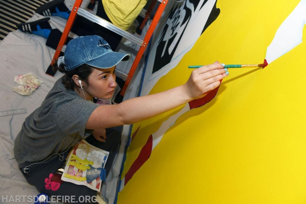 A person with a cap painting a mural on a yellow wall while holding a paintbrush.