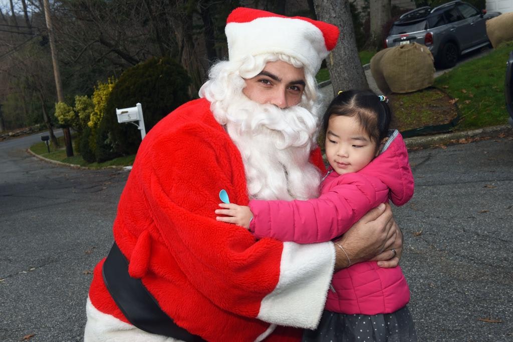 A person in a Santa costume hugs a young child in a pink jacket on a suburban street.