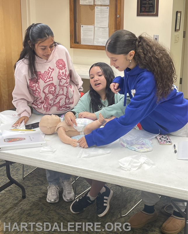 Three young individuals are gathered around a table, interacting with a baby doll and various learning materials.
