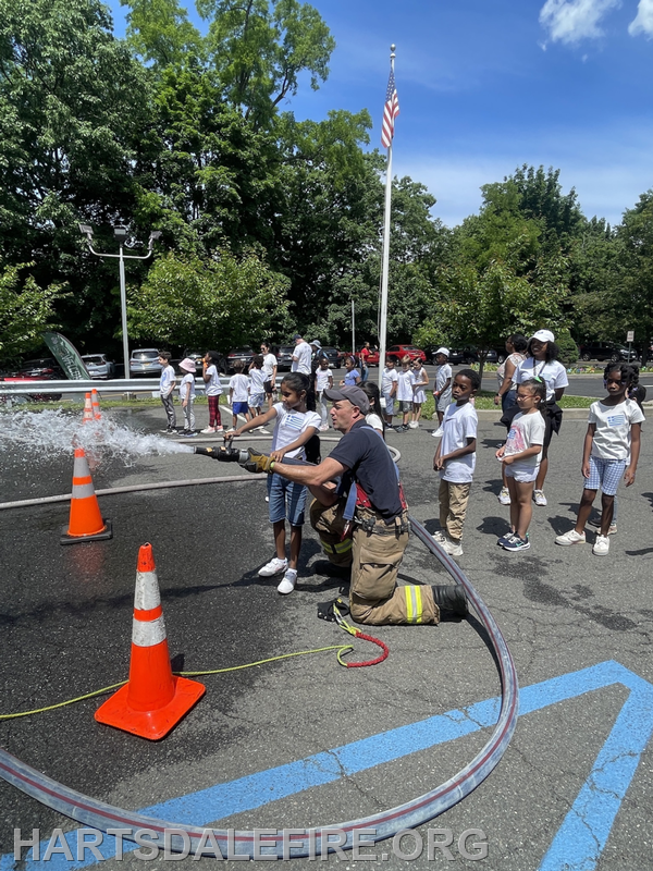 A firefighter demonstrates a water hose to a group of children at a community event, with trees and an American flag in the background.