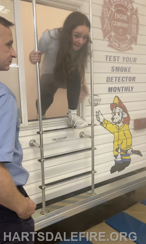 A girl is climbing out of a window, while a man observes nearby; there's a fire safety message on the wall.