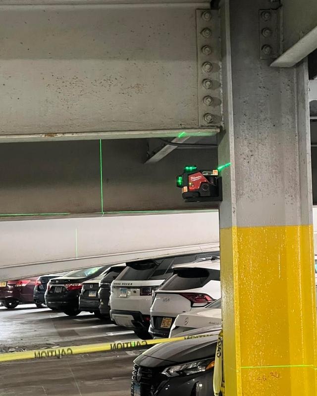 A laser level is mounted on a parking structure beam, projecting green lines, with several parked cars visible in the background.
