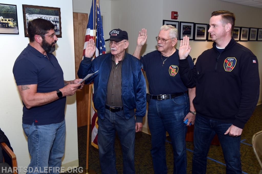 Four men, three taking an oath. Two in fire department attire, one in a cap. An American flag is in the background.