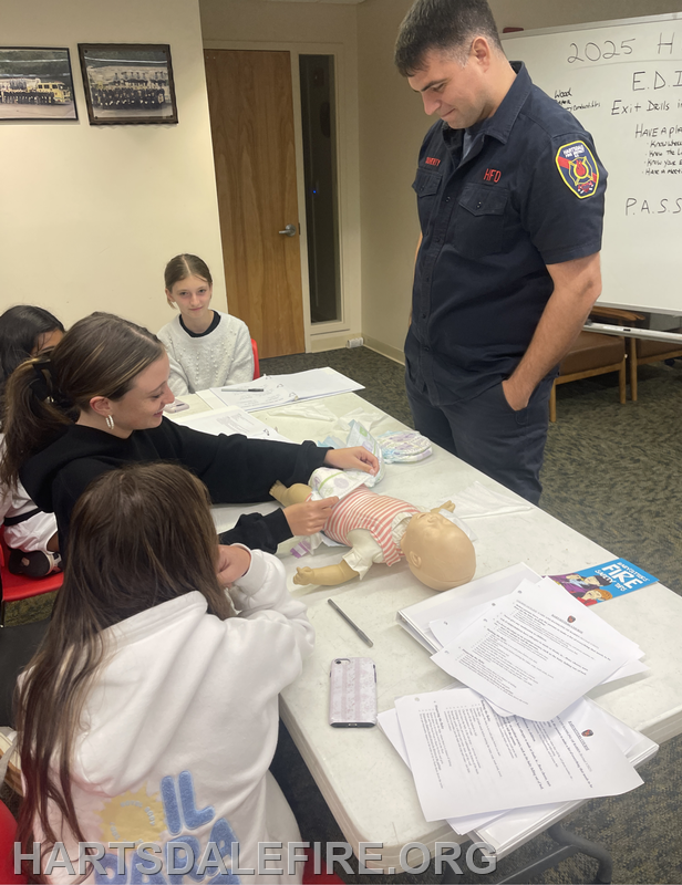 A firefighter teaches a group of young girls how to care for a baby doll, emphasizing safety and caregiving skills.