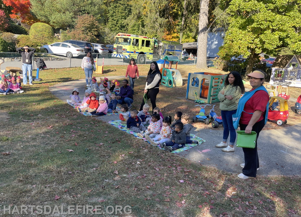 A group of children sits on a mat outdoors with adults nearby, surrounded by play equipment and colorful trees.