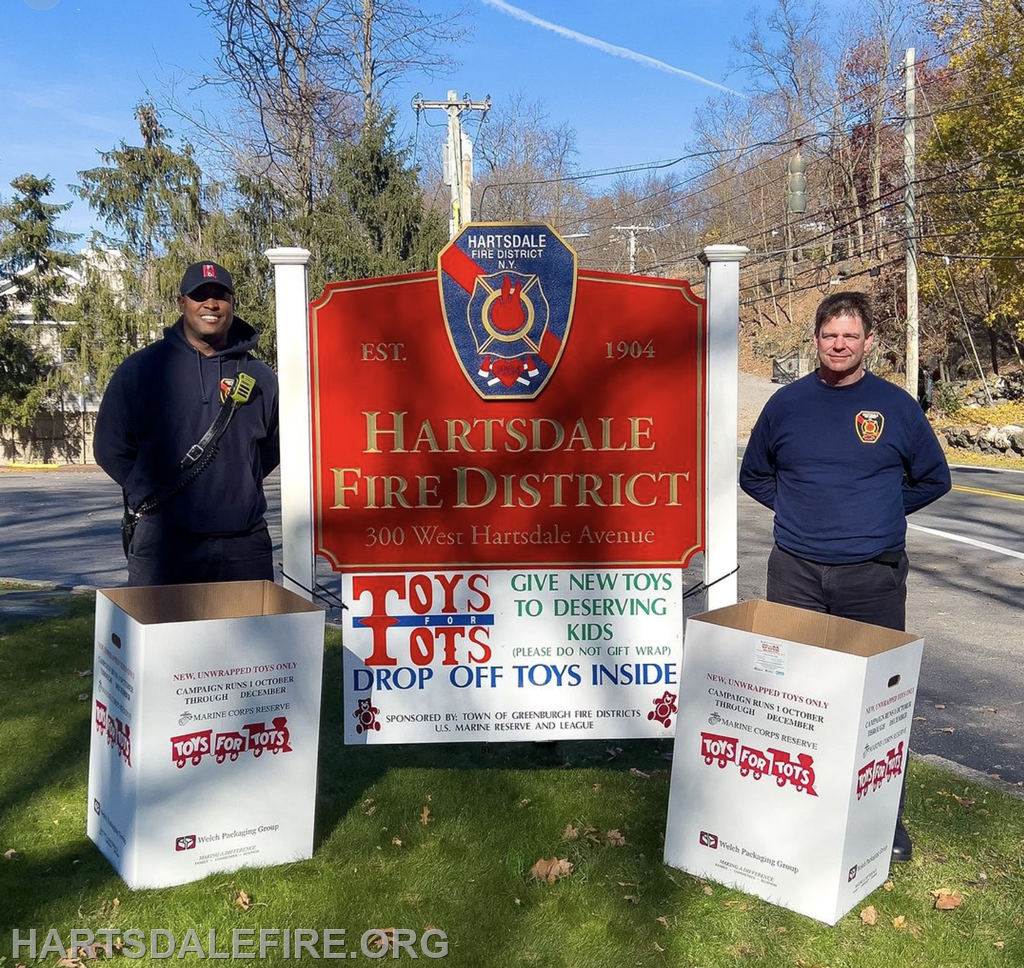 Two firefighters stand by a sign for Hartsdale Fire District, promoting the Toys for Tots donation campaign.