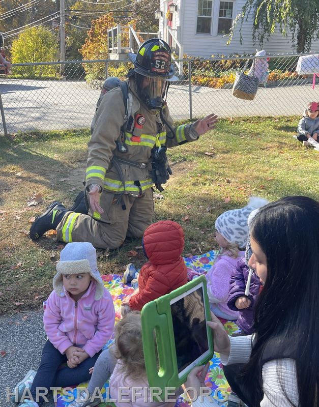 A firefighter kneels while interacting with children sitting on a blanket, with an adult capturing the moment on a tablet.
