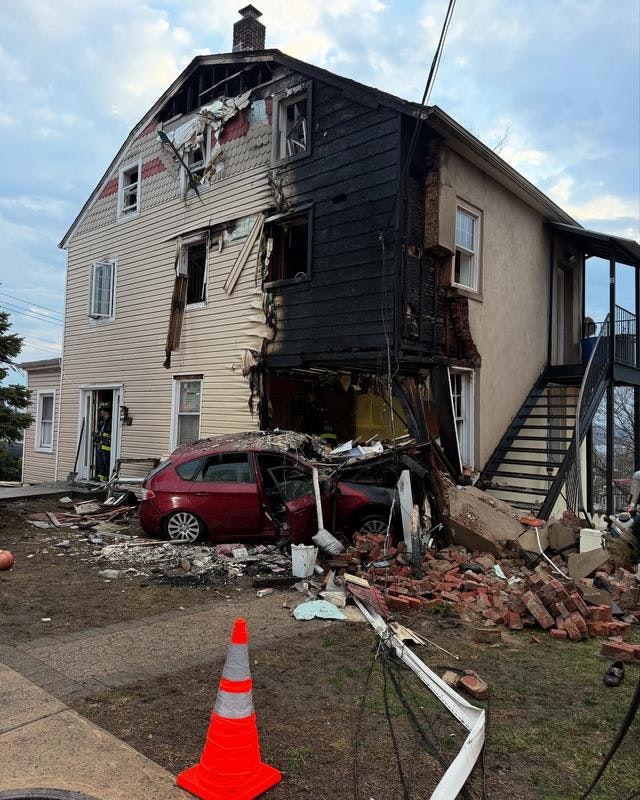 A house with severe damage, partially burned, and a red car crashed into it; debris scattered around and a traffic cone nearby.