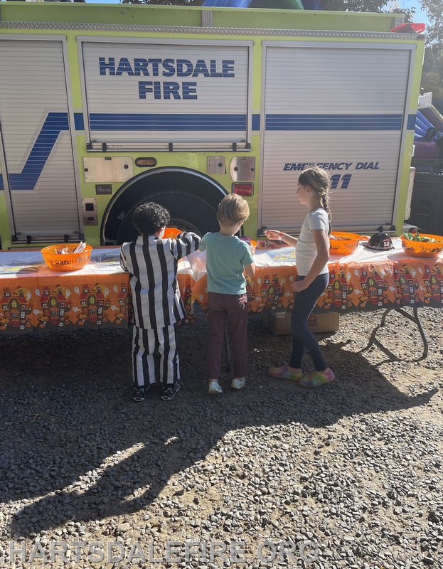 Three children are interacting at a table with Halloween-themed decor near a Hartsdale Fire truck.