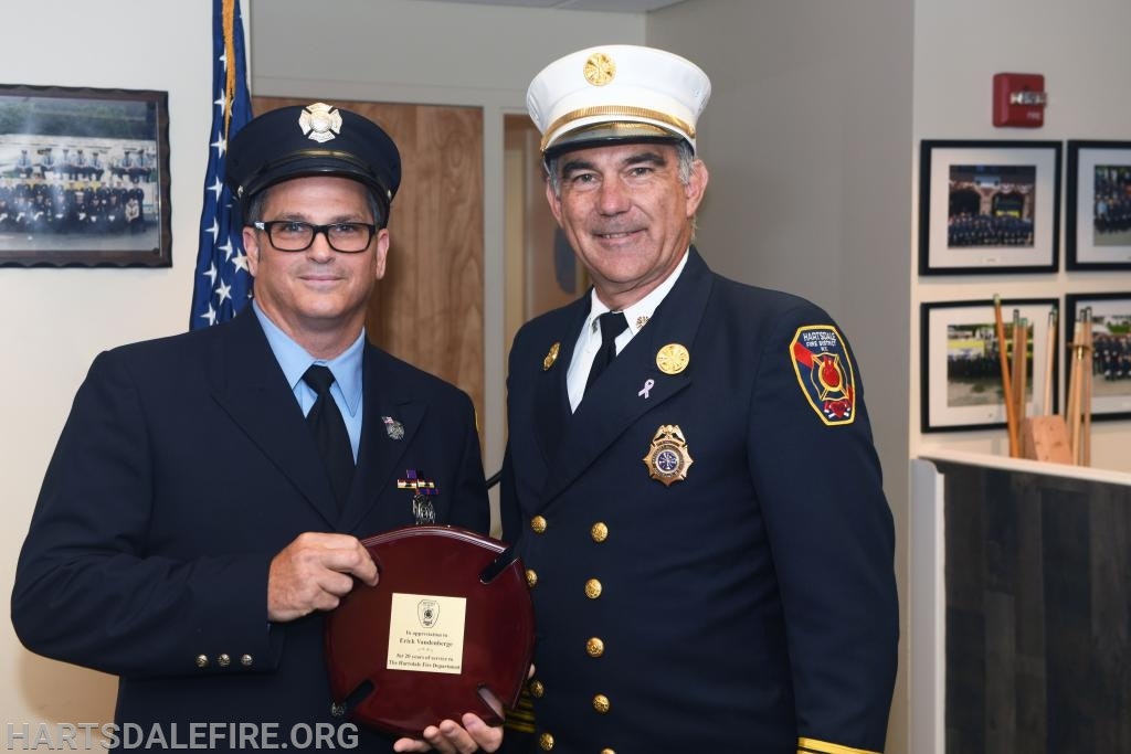 Two men in fire department uniforms holding a plaque in an office setting with framed photos on walls.