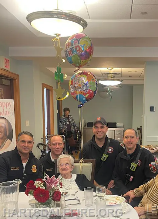 A group celebrates a birthday with balloons at a gathering, featuring an elderly woman and several firefighters.