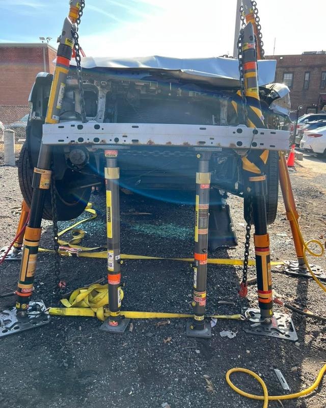 The image shows a car on a supportive frame with hydraulic tools, likely for repairs or inspection.