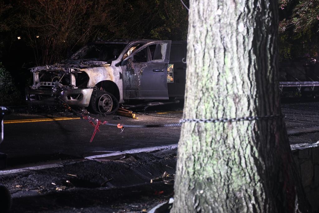 A burned-out vehicle on a road, with its front damaged, near a large tree.