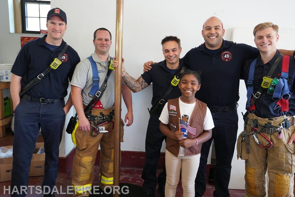 A girl smiles with five firefighters inside a fire station. They are posing together, and some are wearing gear.