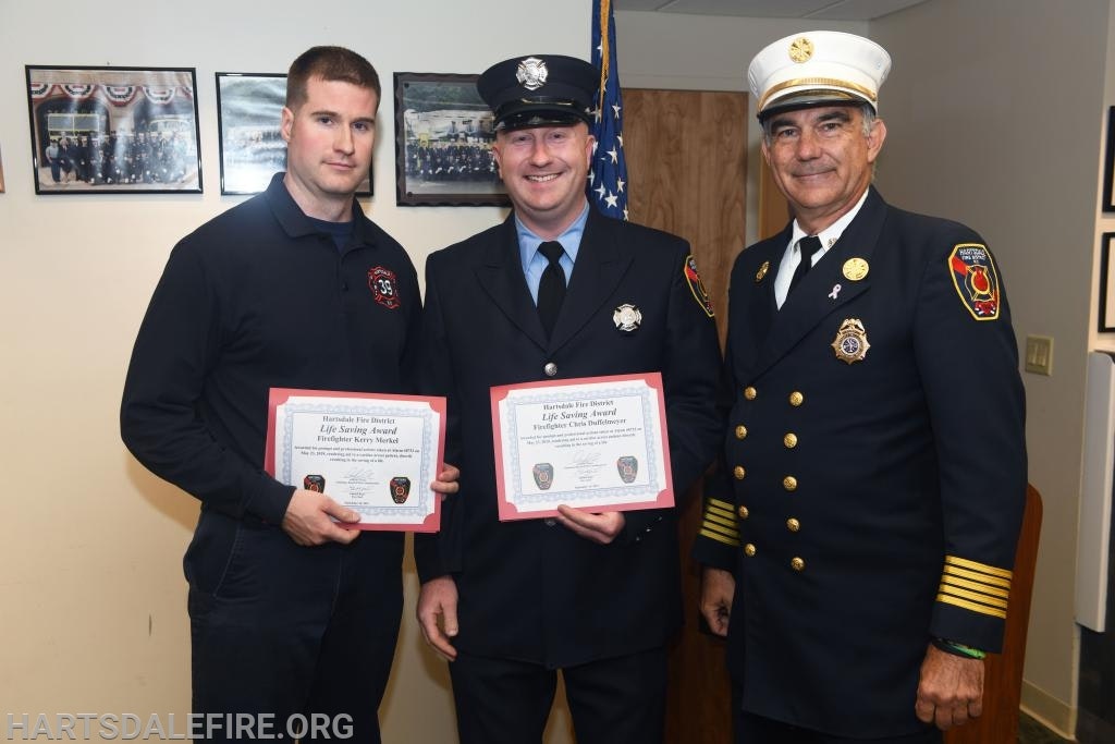 Three firefighters in uniform, two holding certificates, smiling in a room with framed photos on the wall.