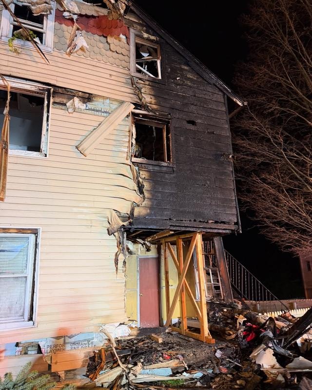 A partially burned house shows extensive damage, with charred siding and debris around the base, indicating a recent fire.