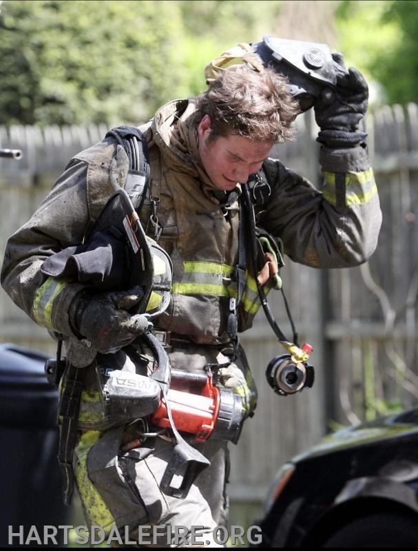 A firefighter in gear holds his helmet, appearing tired, with fire equipment and the website "HARTSDALEFIRE.ORG" visible.