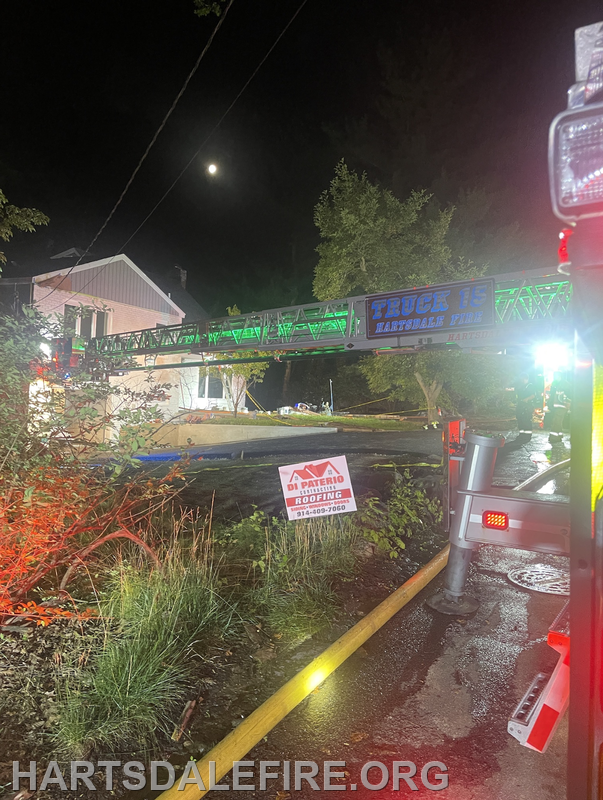 A fire truck's ladder is extended at night near a house, with emergency lights and a "Roofing" sign visible in the foreground.