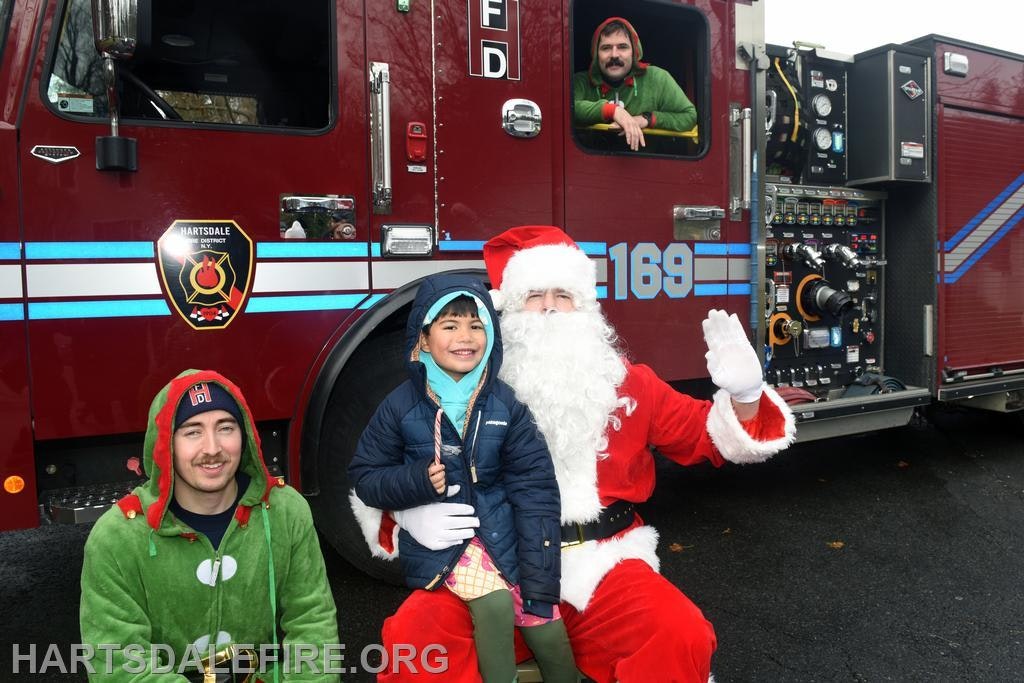 A festive scene with Santa, a child, and two people in holiday attire by a fire truck, spreading holiday cheer.