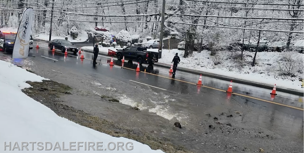 The image shows a snow-covered street with police vehicles and traffic cones, indicating a possible road closure due to water issues.
