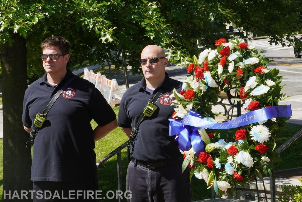 Two fire department members stand near a floral wreath, likely commemorating a special event or memorial.