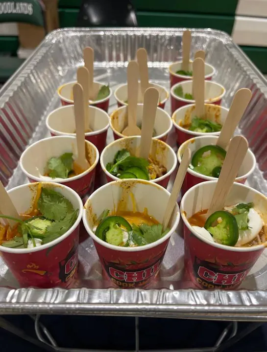 A tray of small cups filled with chili, garnished with cilantro and jalapeño slices, each with a wooden stick for sampling.
