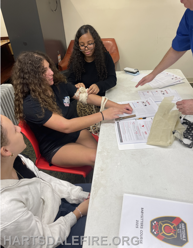 Participants in a babysitting course practice first aid skills, like applying a bandage, at a table with instructional materials.