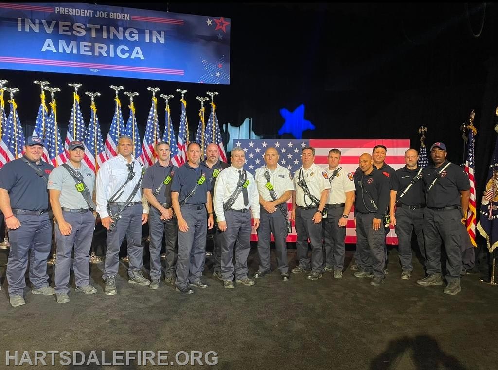 A group of firefighters stands in front of a U.S. flag and "Investing in America" sign with multiple American flags behind them.