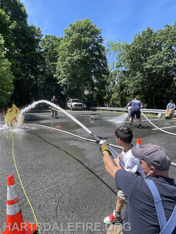 A child and adult spray water from a fire hose at a training exercise, with fire safety equipment and trees in the background.