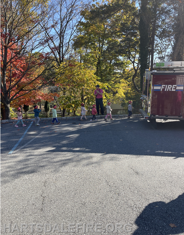 A group of children in hats walks near a fire truck, with colorful trees in the background on a sunny day.