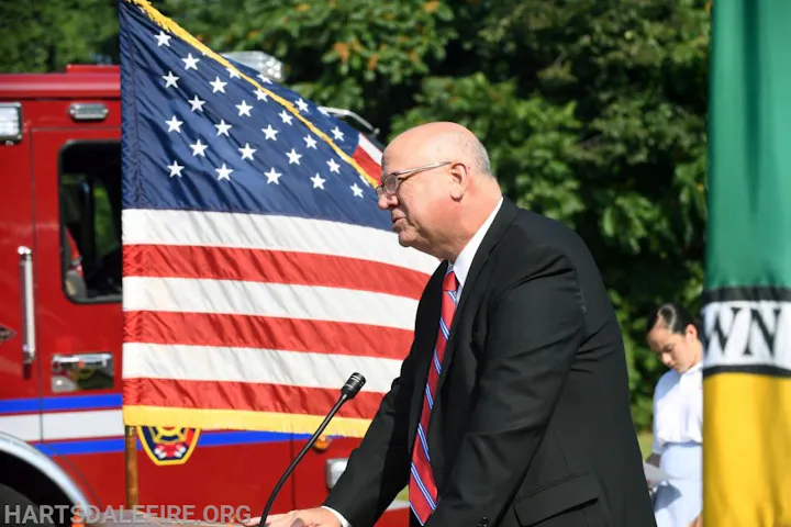 A man in a suit speaks at a podium, with a U.S. flag and fire truck in the background.