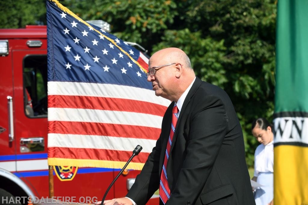 A man in a suit speaks at a podium, with a U.S. flag and fire truck in the background.