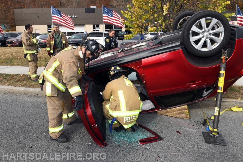 Firefighters are responding to an overturned car, assisting inside while onlookers watch nearby. American flags are visible.